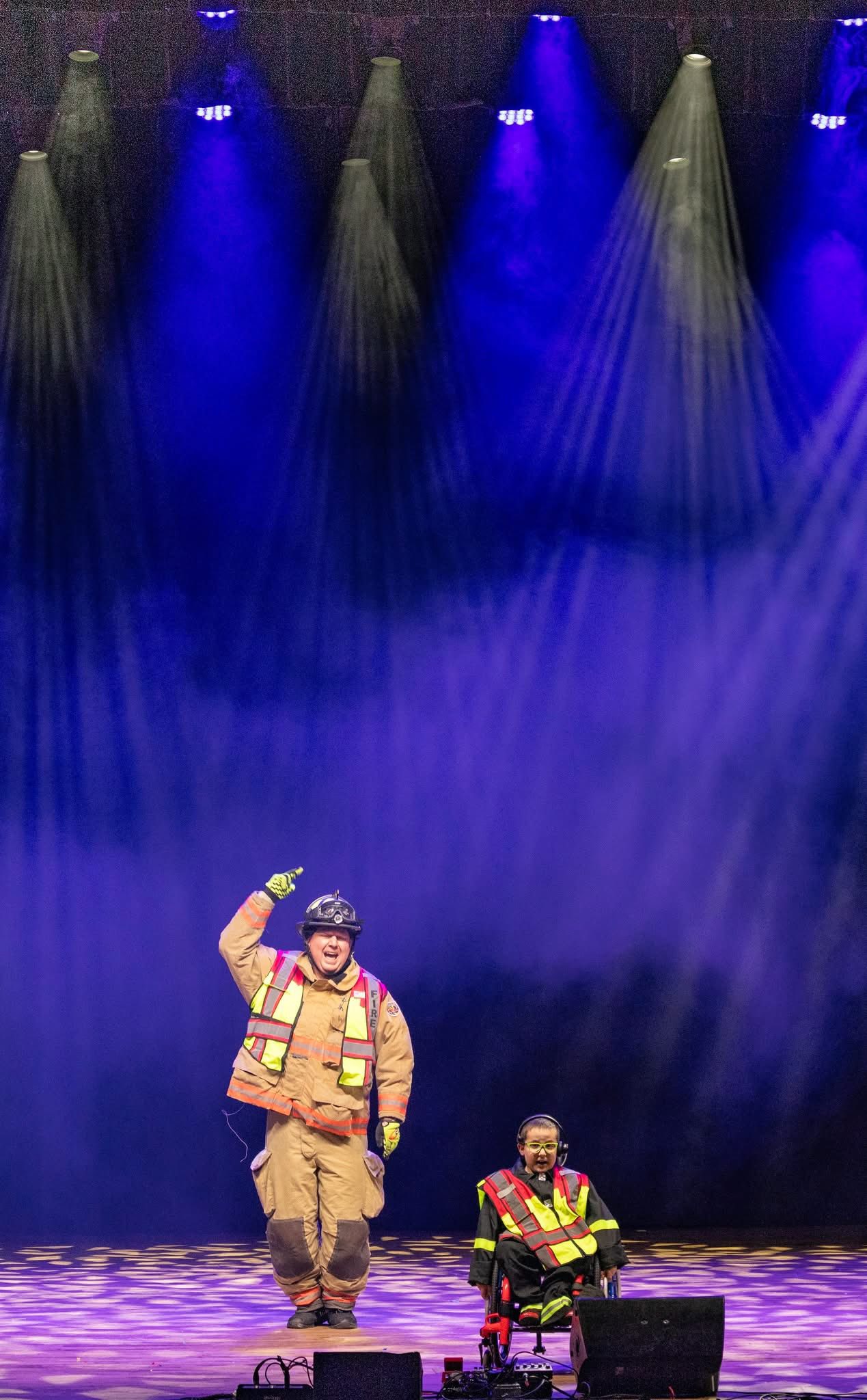 Two performers dressed as firefighters, one standing and pointing up, the other in a wheelchair, on a stage with blue and purple spotlights at the Lip Sync Battle at at The Paramount Theatre.
