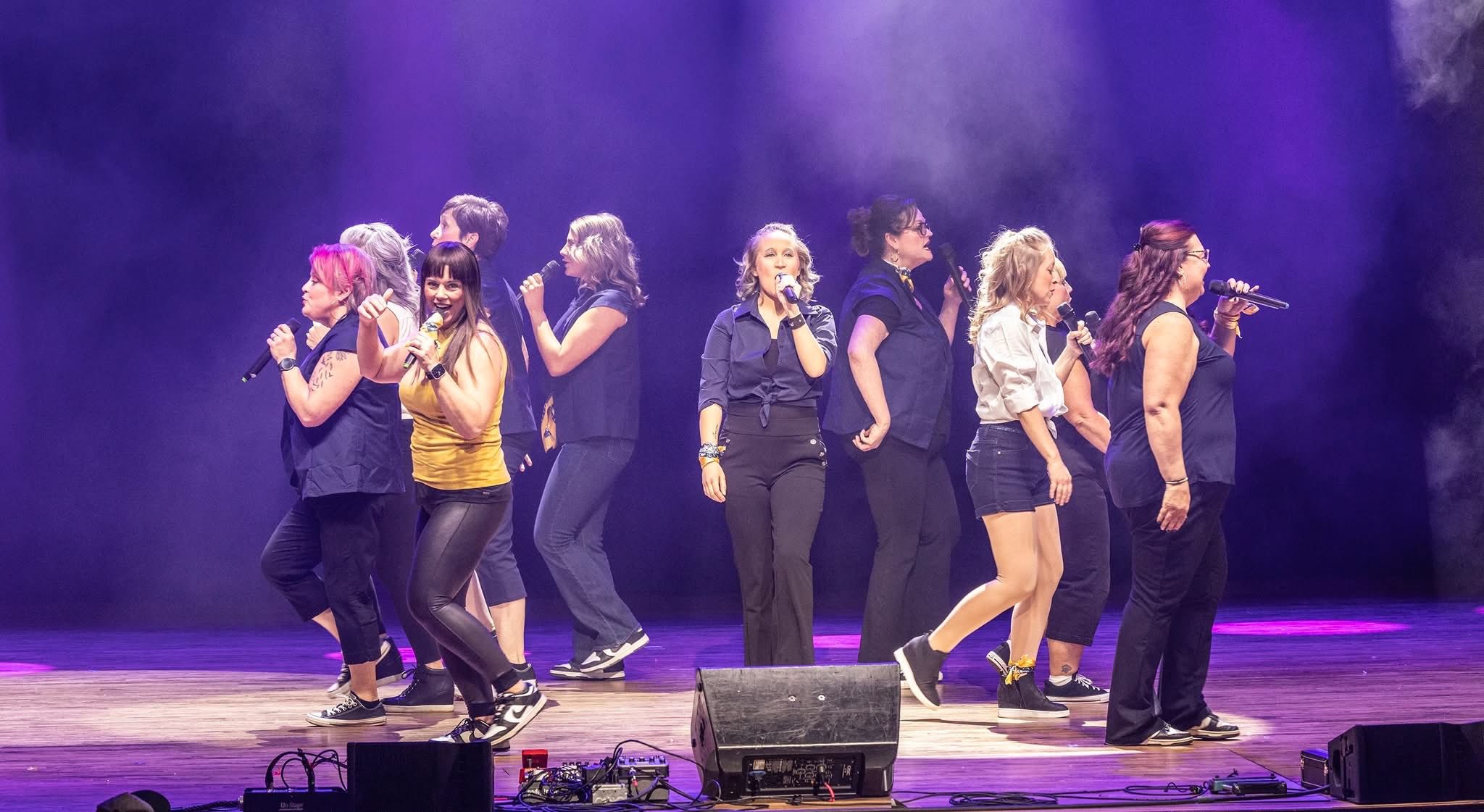 Group of ten women singing on stage with microphones under purple lighting and slight fog at the Lip Sync Battle at at The Paramount Theatre.