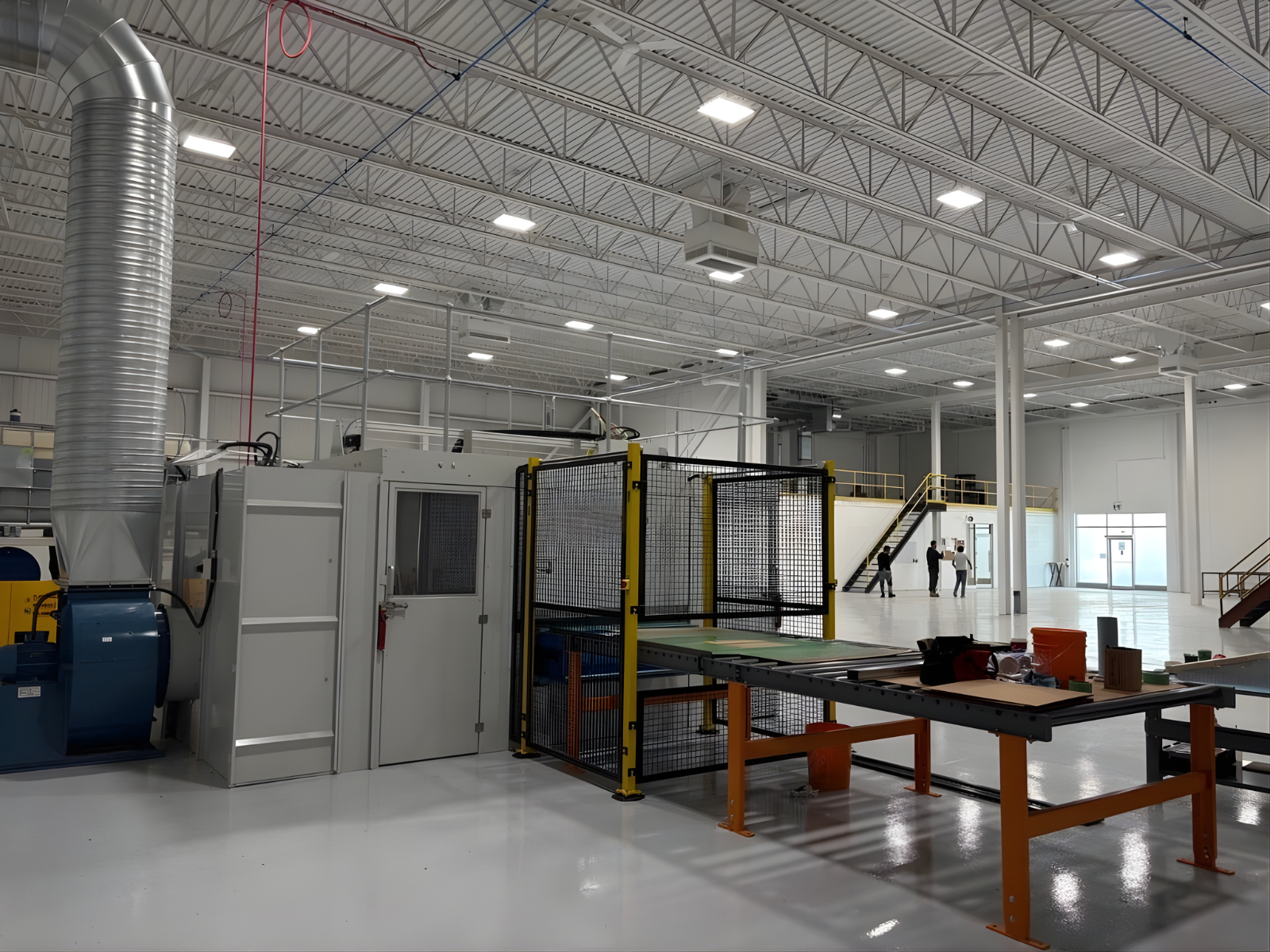 Spacious industrial warehouse with machinery, safety cages, and three people talking near a staircase.
