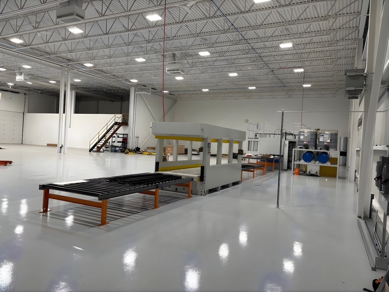 Spacious, well-lit industrial warehouse with machinery, metal roller conveyor, and storage containers.