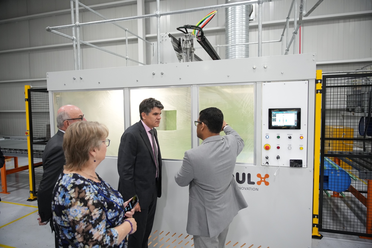 Four professionals in business attire standing near a large industrial machine with a control panel in a factory setting.