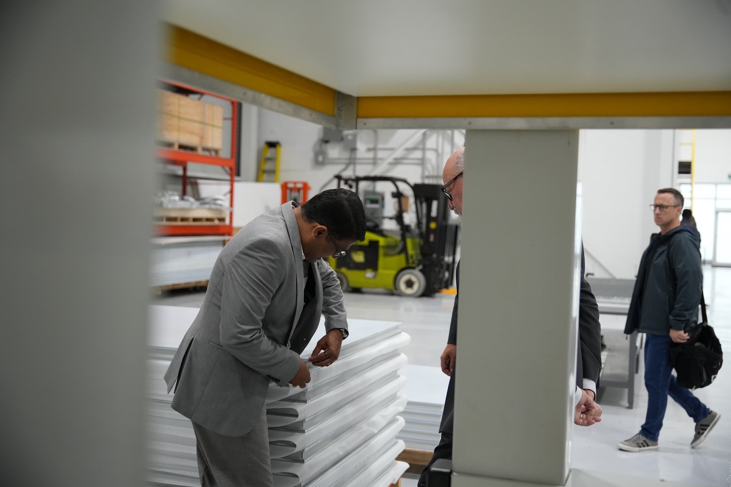 Man in a gray suit closely inspecting stacked white materials in a warehouse while another man in a suit and a casually dressed man walk nearby.