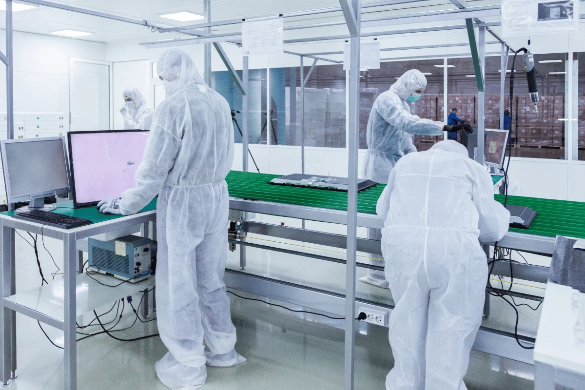 Laboratory workers in white protective suits operate computers and equipment on a production line in a sterile, high-tech, cleanroom environment.