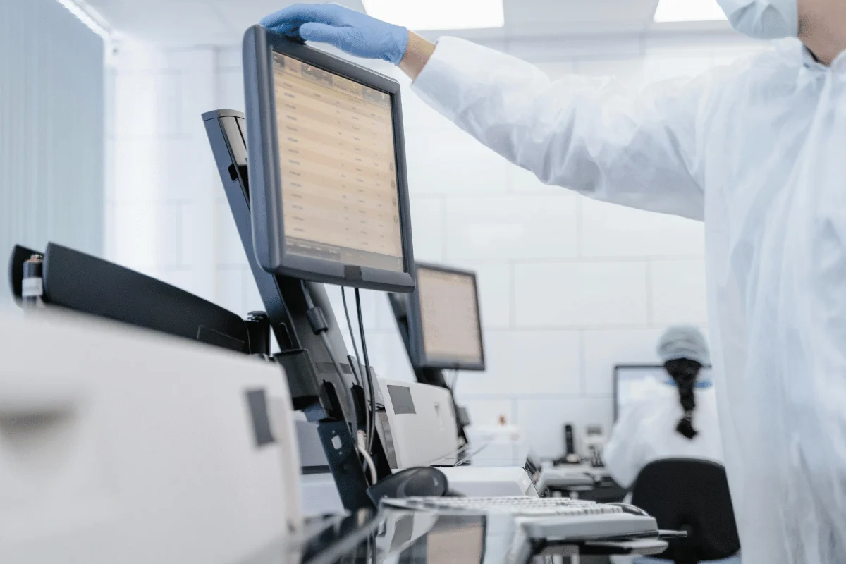 Scientist in a lab coat and gloves adjusts a monitor, surrounded by computers and lab equipment, in a clean, sterile laboratory environment.