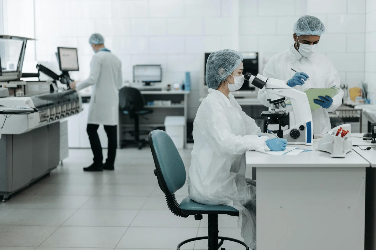 A laboratory scene with two scientists in protective gear, one using a microscope and the other taking notes at a desk.