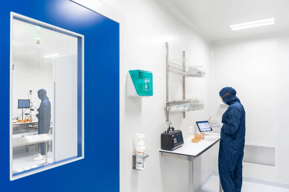 A person in a blue lab coat and mask works with a tablet in a sterile lab, surrounded by shelves and petri dishes.
