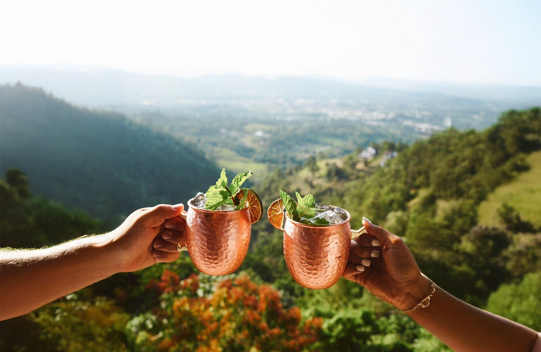 Two hands cheering with drinks with Jarabacoa in the background.