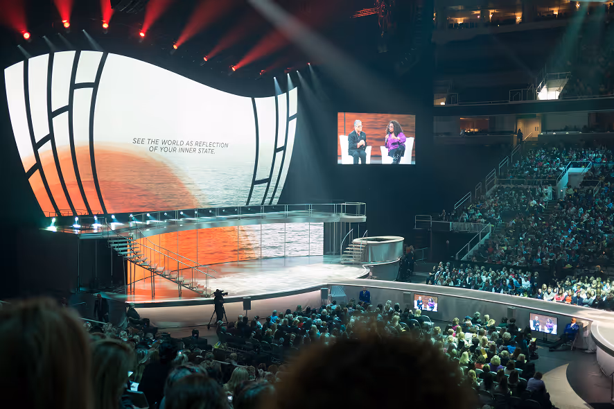 Large indoor arena filled with audience watching a stage with bright curved screens displaying a sunset over water and a message.