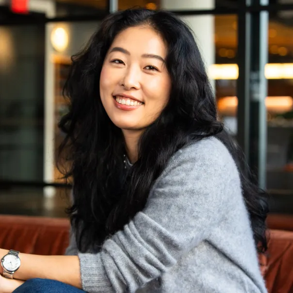 Smiling woman with long black hair wearing a gray sweater, sitting indoors.