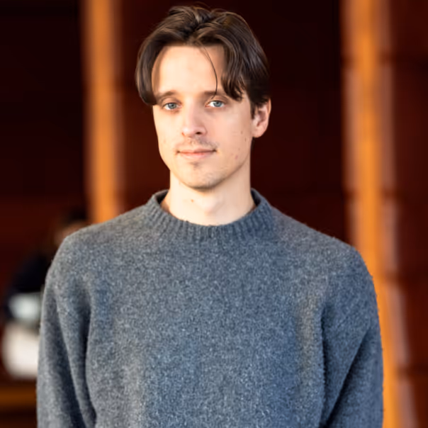 Young man with light skin and brown hair wearing a gray sweater, standing indoors with a blurred warm-toned background.