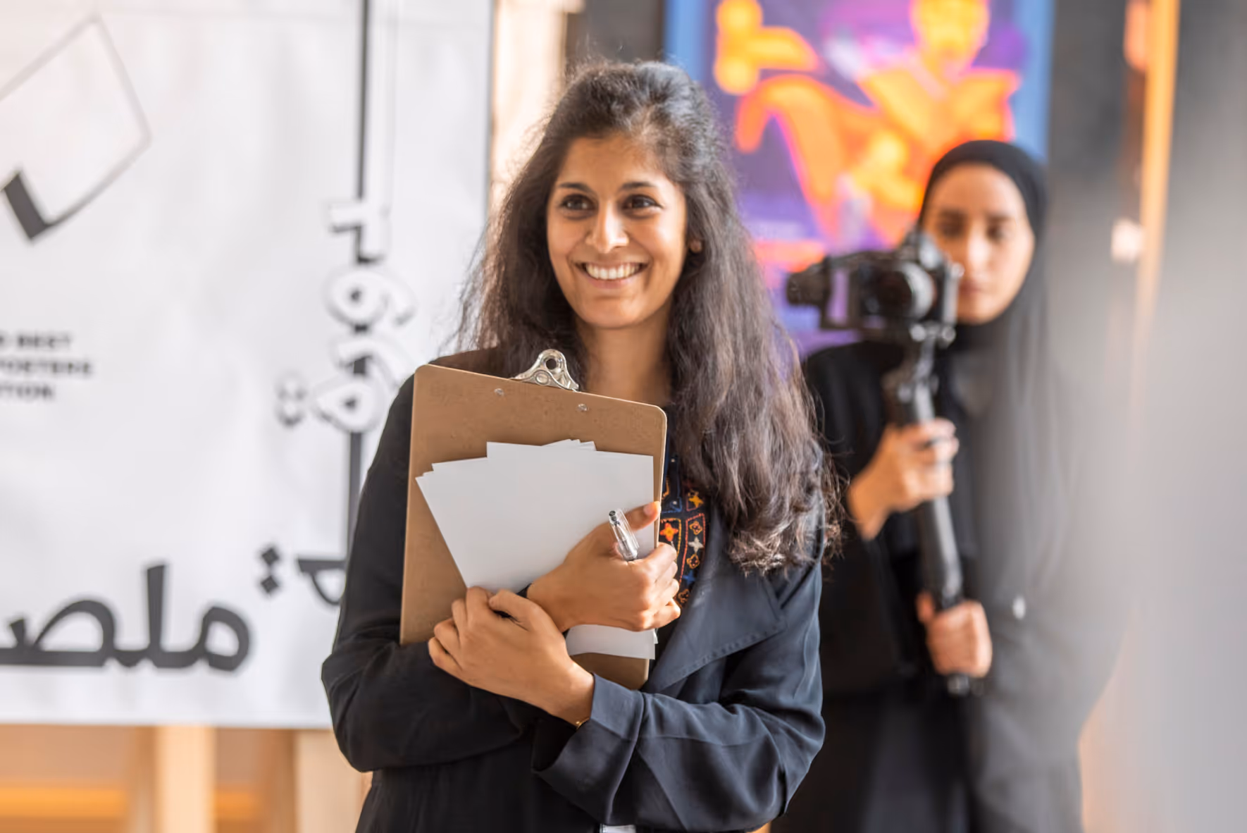 Smiling woman holding a clipboard with papers and a pen, with another woman in a hijab holding a camera in the background.