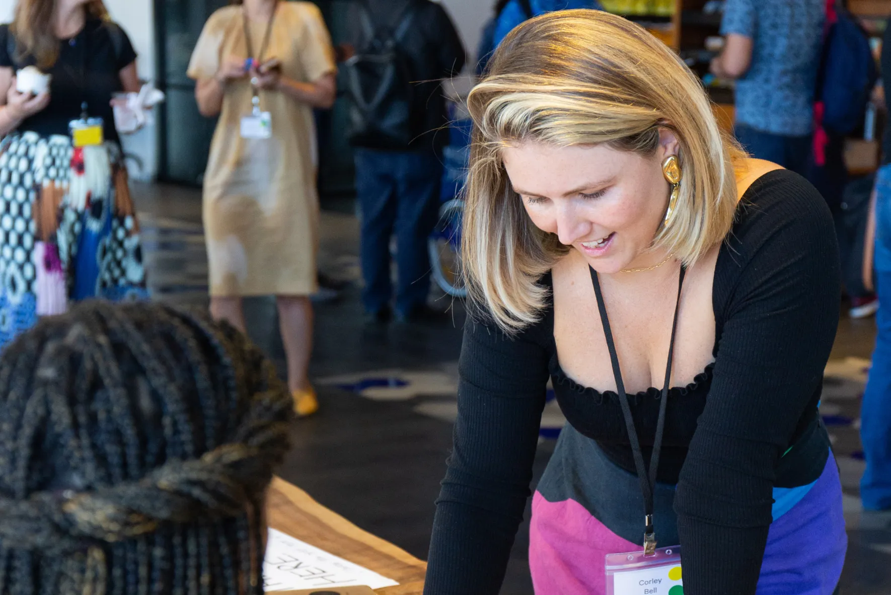 Blonde woman with name badge leaning forward and talking to a person with braided hair at a table during an event.