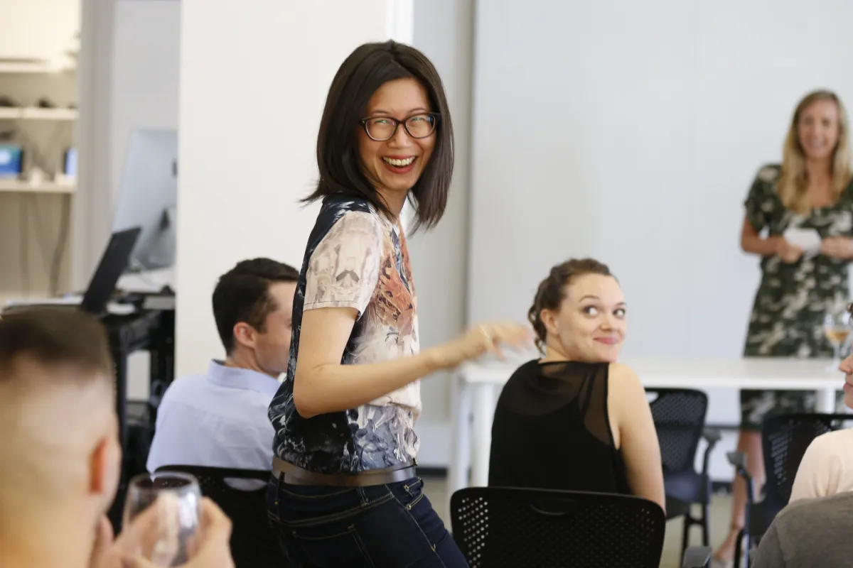 Smiling woman standing and turning toward the camera in a casual office meeting with colleagues seated around.