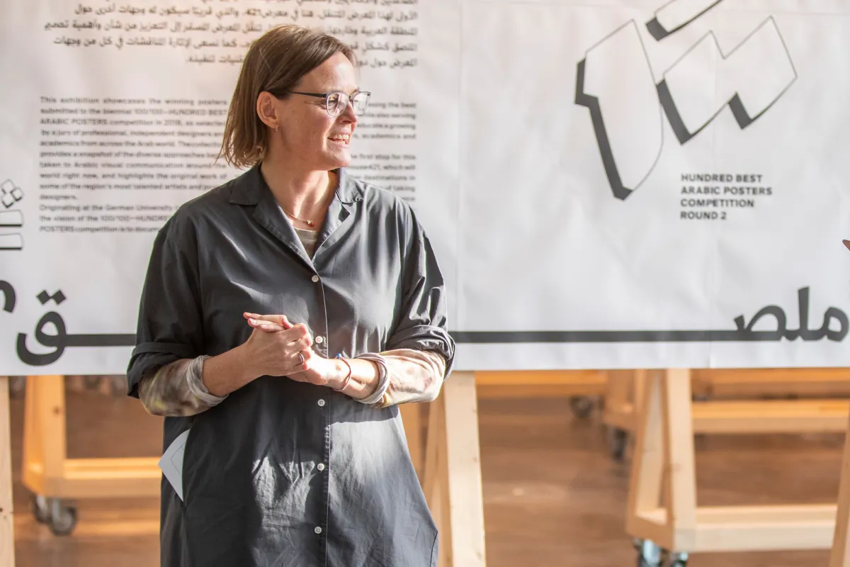 Woman with glasses smiling and standing in front of a poster for the Hundred Best Arabic Posters Competition Round 2.