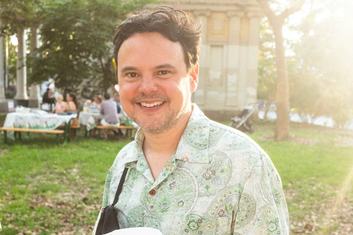 Smiling man with curly hair wearing a patterned light green shirt outdoors in a sunny park setting.