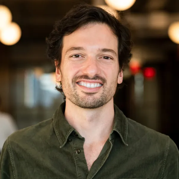 Smiling man with dark hair and beard wearing a green button-up shirt in a softly lit indoor setting.