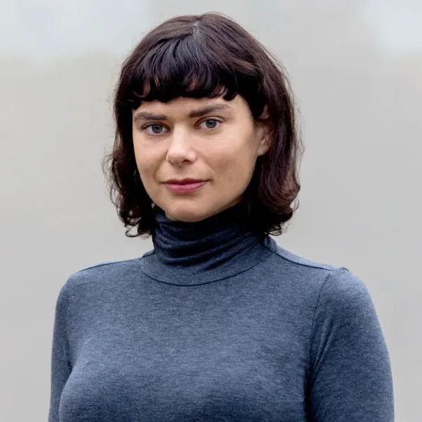 Portrait of a woman with short curly dark hair and bangs, wearing a gray turtleneck sweater against a plain light background.