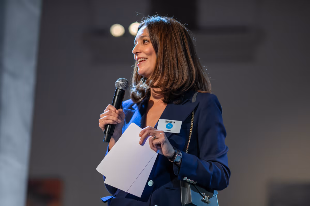 Woman in blue blazer holding a microphone and papers, speaking and smiling.