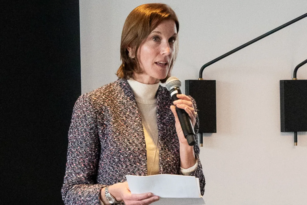 Woman with brown hair speaking into a microphone while holding a paper, wearing a patterned blazer and white turtleneck.
