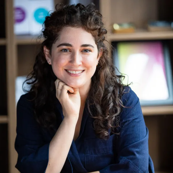 Smiling woman with curly brown hair wearing a navy blue shirt, resting her chin on her hand in a cozy room with wooden shelves.