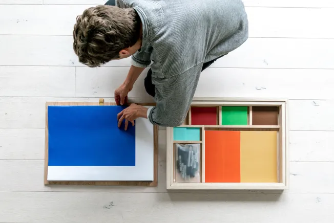 Overhead view of a person organizing colorful paper sheets and markers on a white wooden floor.