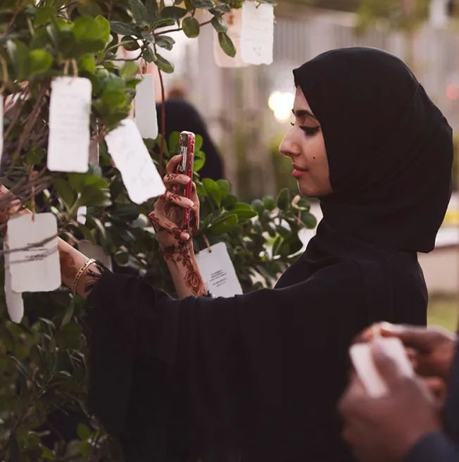 Woman wearing a black hijab and henna designs on her hand, holding a smartphone and reading notes hanging on a leafy tree.