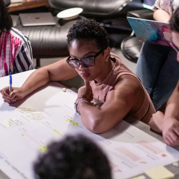 Person wearing glasses writing notes on a large white poster paper with others around a table in a meeting.