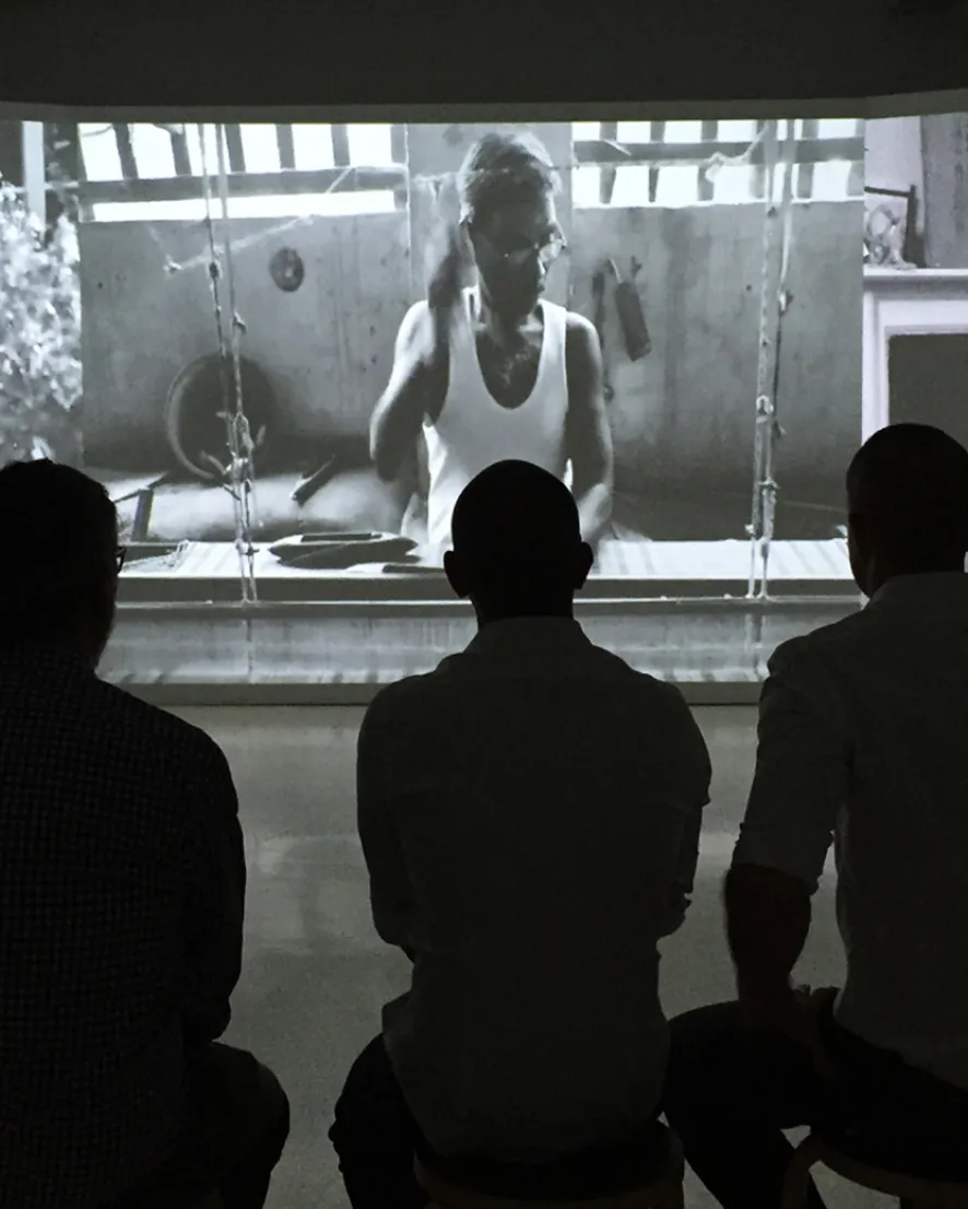 Three men sitting in silhouette watching a black-and-white film of a person working with tools indoors.
