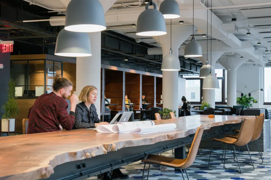 Two people working together at a large wooden table in a modern, open office space with hanging lights and several people in the background.
