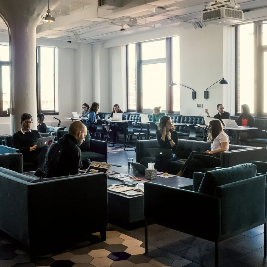 Modern co-working space with people working on laptops and engaging in conversation on sofas and at tables by large windows.