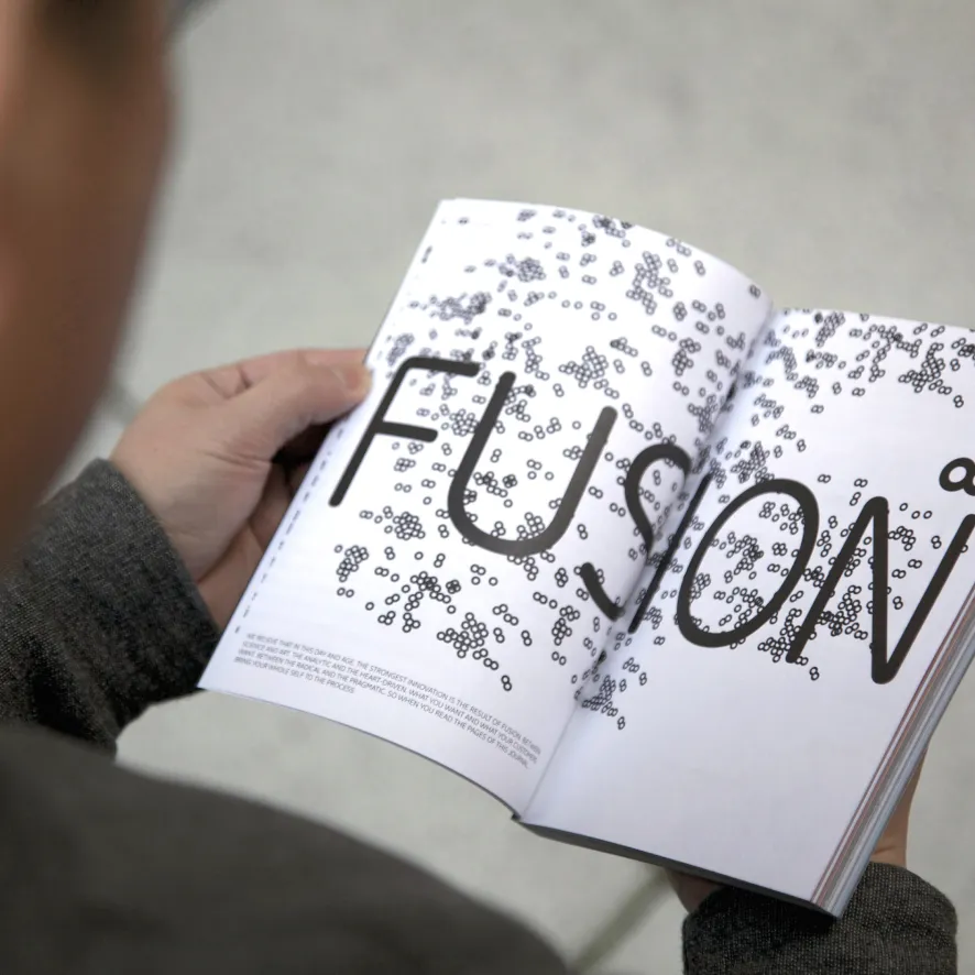Person holding and reading an open book with the word 'Fusion' in large letters and scattered black dot patterns on white pages.
