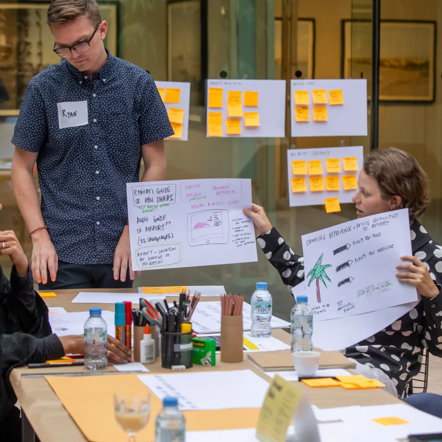 Three people at a table in a meeting room with sticky notes and papers; one man wearing glasses and a name tag 'Ryan' stands holding a paper with notes while a woman seated holds another paper discussing ideas.