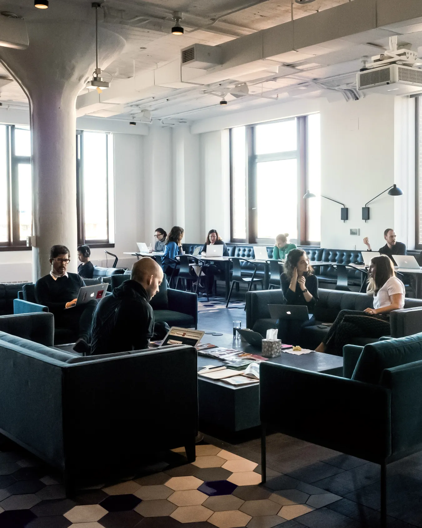 Modern coworking space with people working on laptops and conversing on sofas and chairs near large windows.