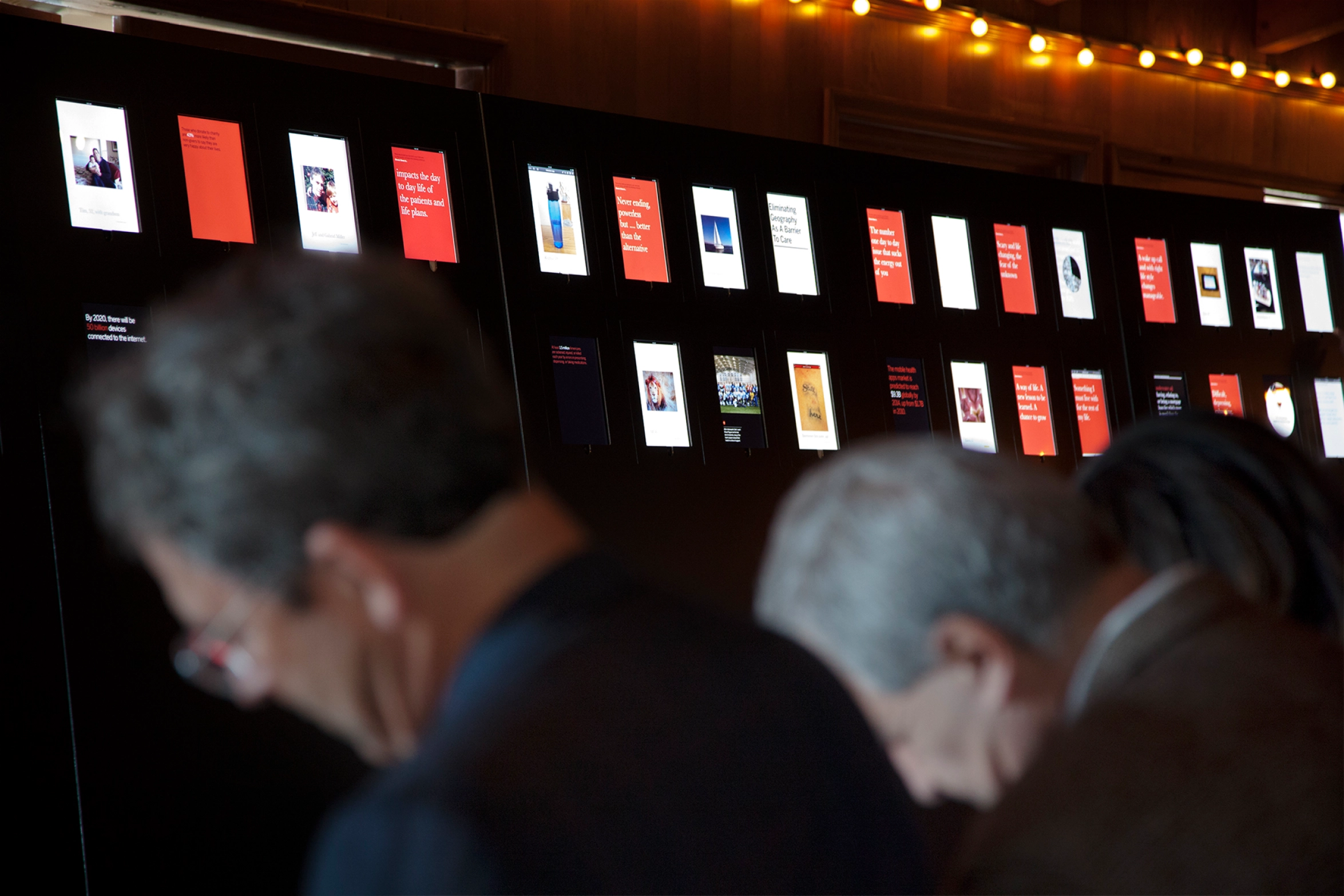 Two elderly people with heads bowed in front of a large digital display wall showing various images and text panels.