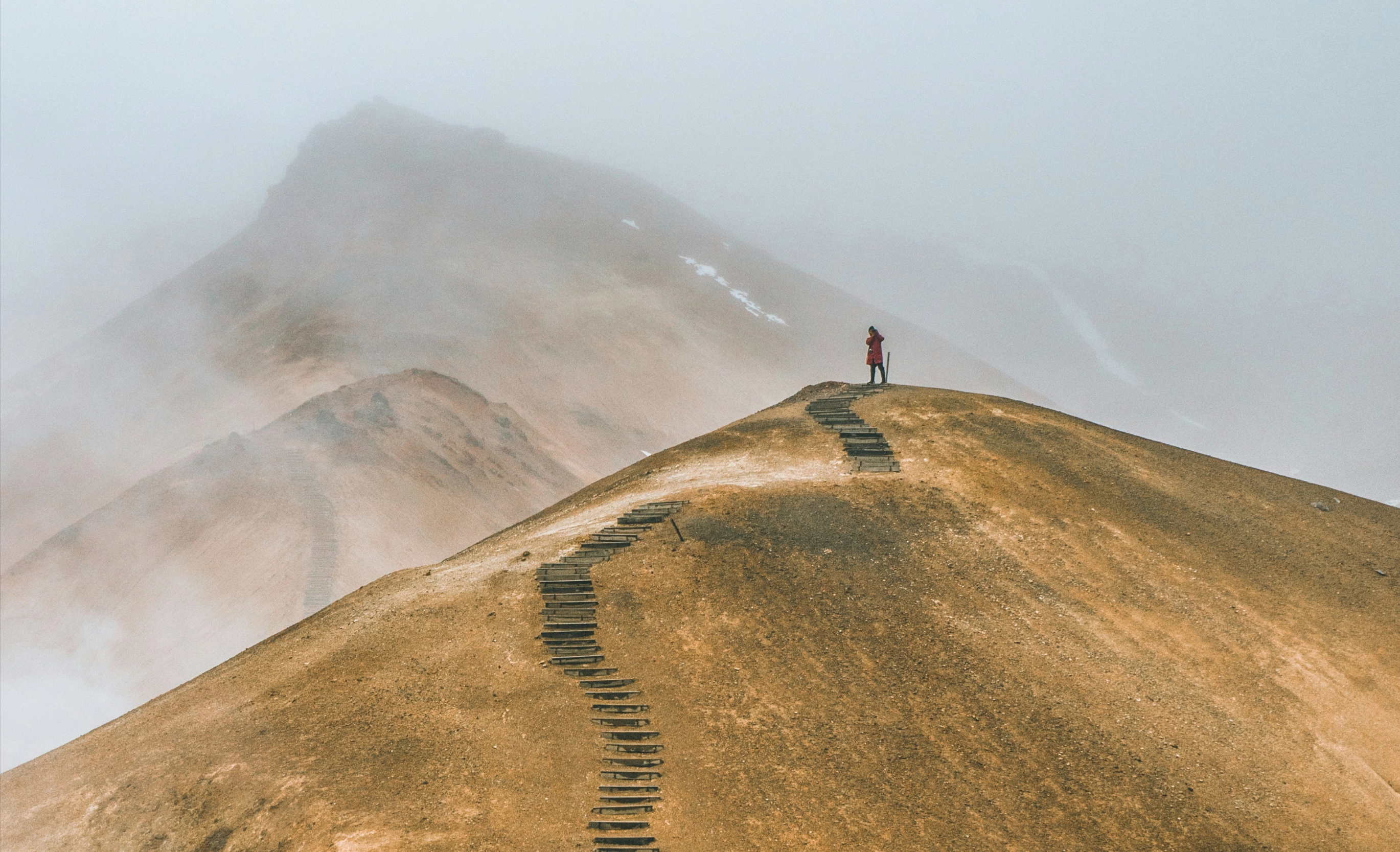Person in red coat standing on top of a foggy, barren mountain with wooden steps leading up.