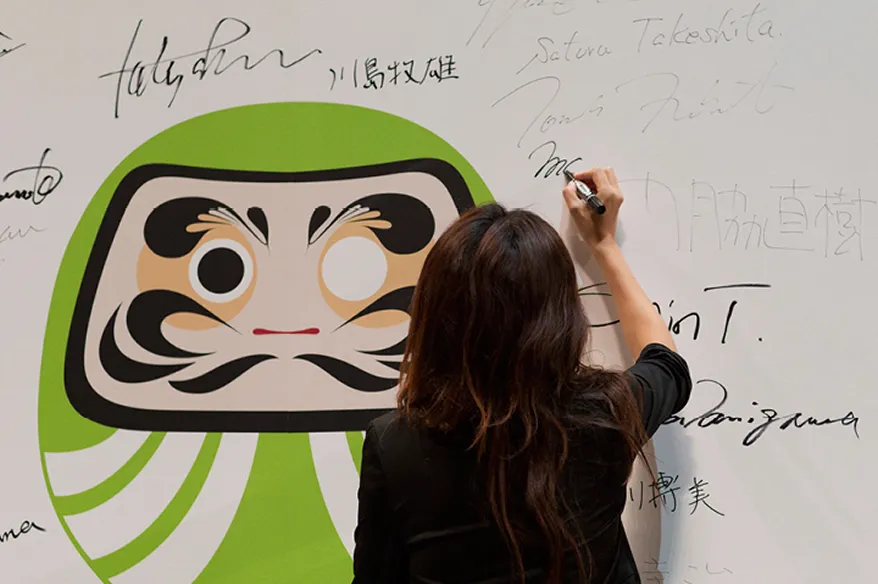 Person with long hair signing a white wall next to a large green Daruma doll illustration with one eye painted.