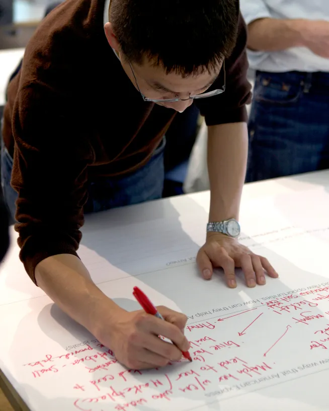 Person wearing glasses and a brown sweater writing notes with a red marker on a large white sheet of paper.