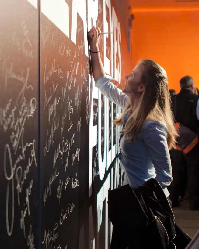 Smiling woman signing a large black wall filled with handwritten signatures, holding a jacket in her left hand.