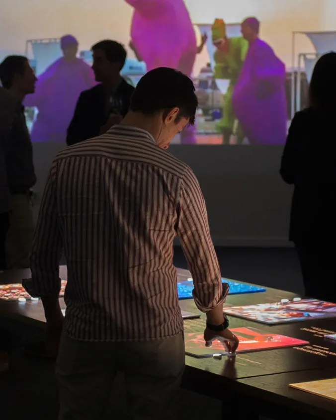 Person in a striped shirt interacting with illuminated panels on a table at an exhibit with a colorful projection in the background.