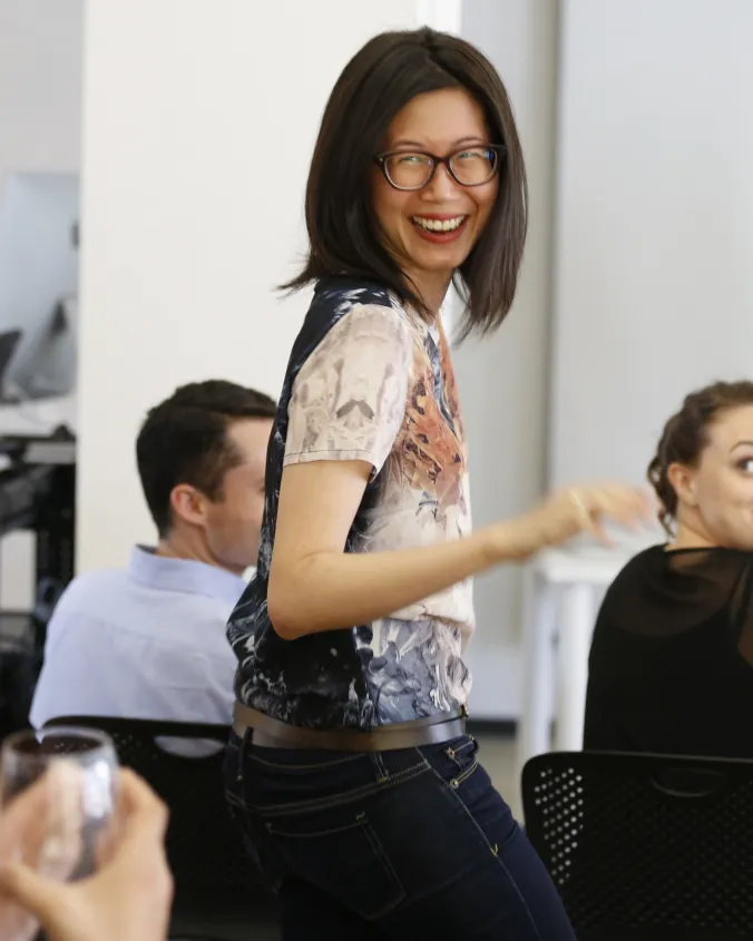 Smiling woman with glasses wearing a colorful shirt and jeans standing in an office environment with seated colleagues.