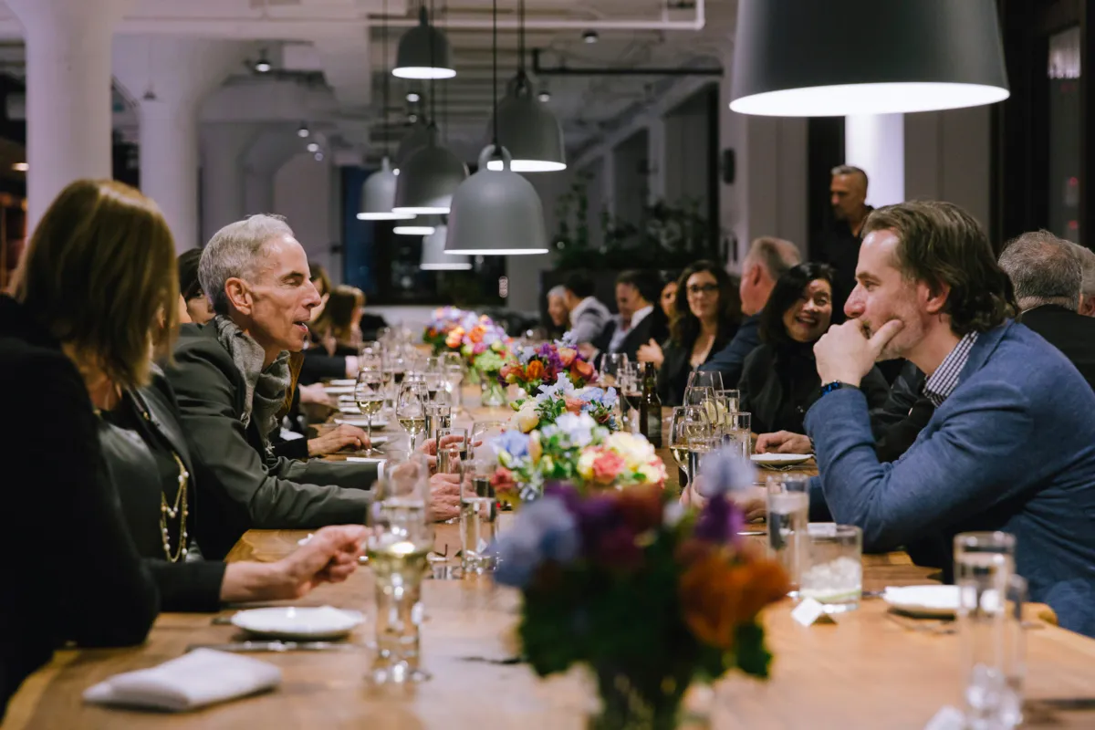 People sitting and conversing at a long dining table decorated with colorful flower arrangements and glassware in a modern indoor setting.