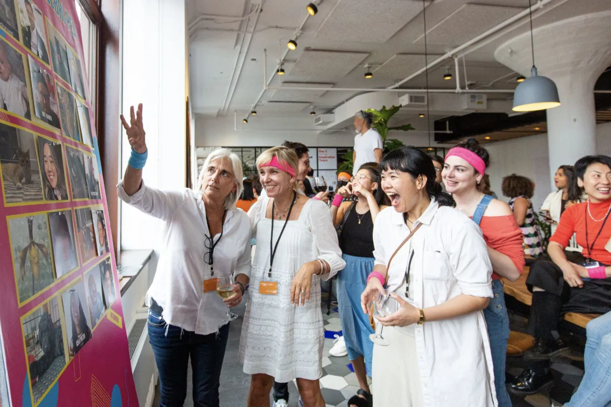 A group of diverse people indoors looking excitedly at a large photo board, with some wearing pink headbands and wristbands and holding drinks.