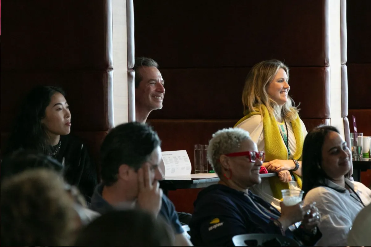 A diverse group of people sitting and smiling attentively in a dimly lit room with dark brown walls.
