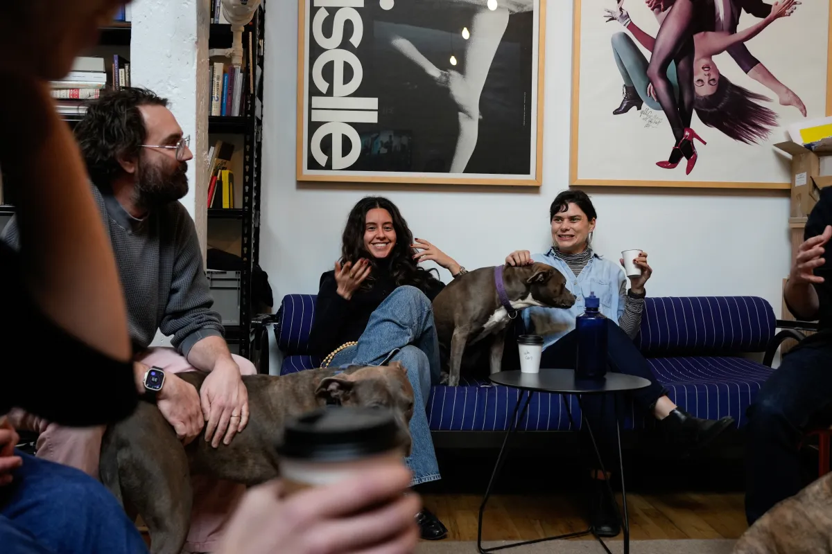 Group of people sitting in a cozy room with two dogs, smiling and holding coffee cups.