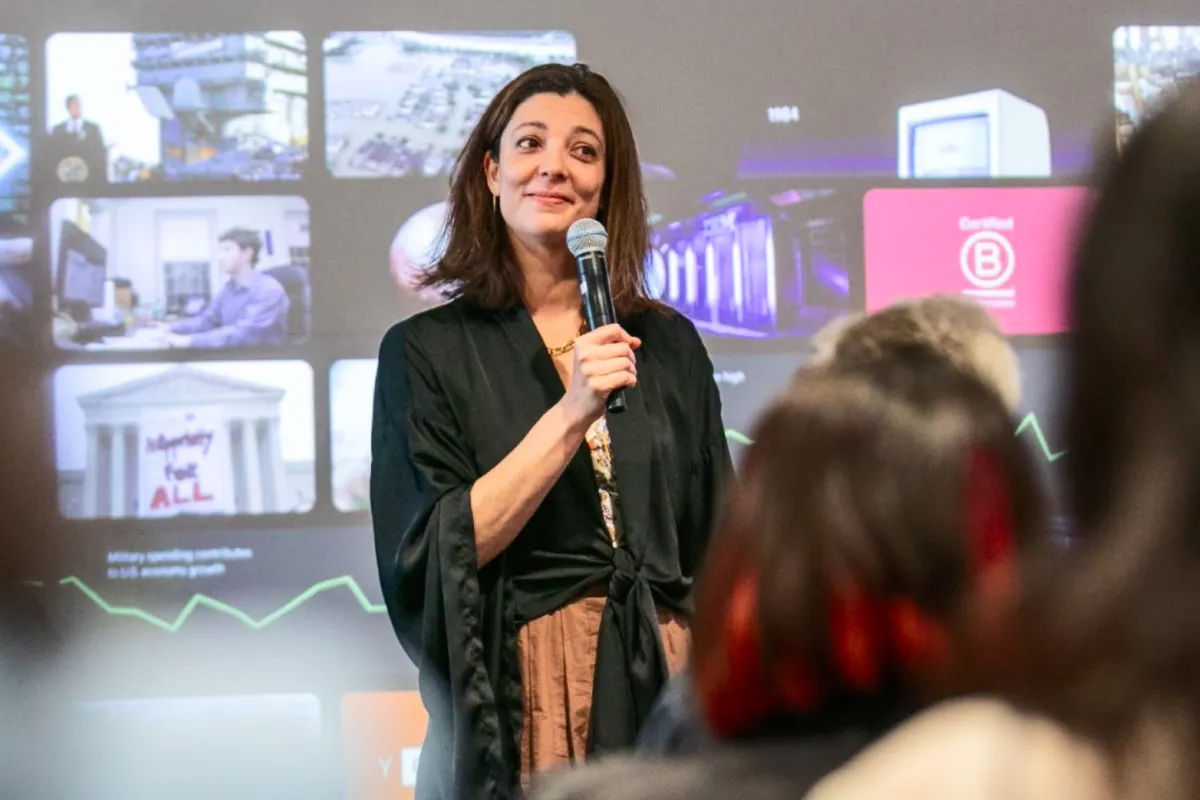 Woman speaking into a microphone in front of a screen displaying various images and data to an audience.