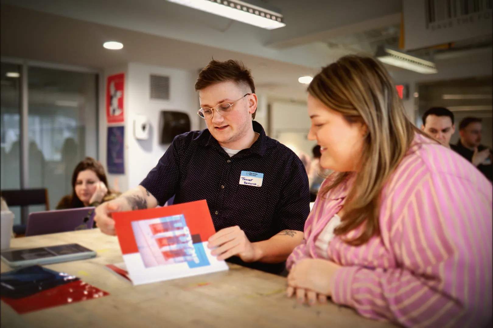 A man wearing glasses and a name tag shows a colorful booklet to a smiling woman in a pink striped shirt at a table.