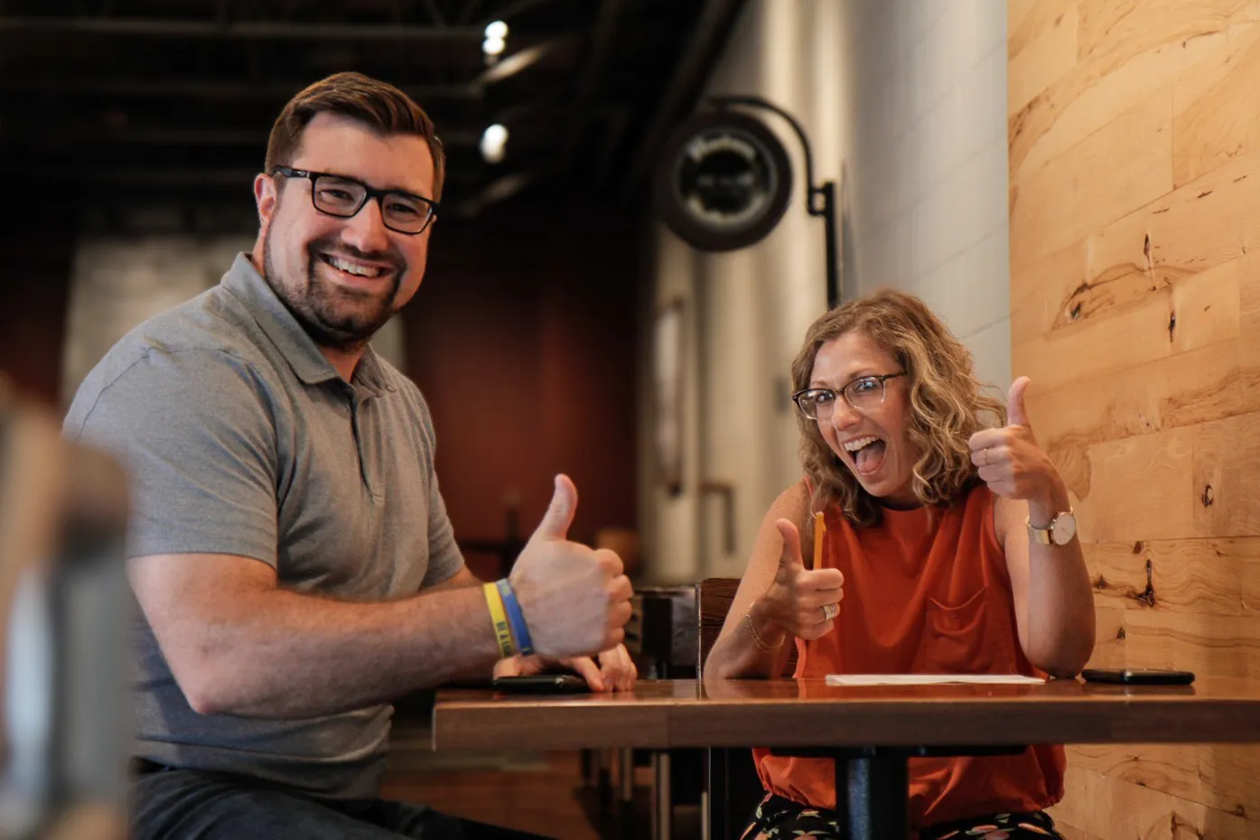 Man and woman sitting at a table smiling and giving thumbs up in a casual indoor setting.