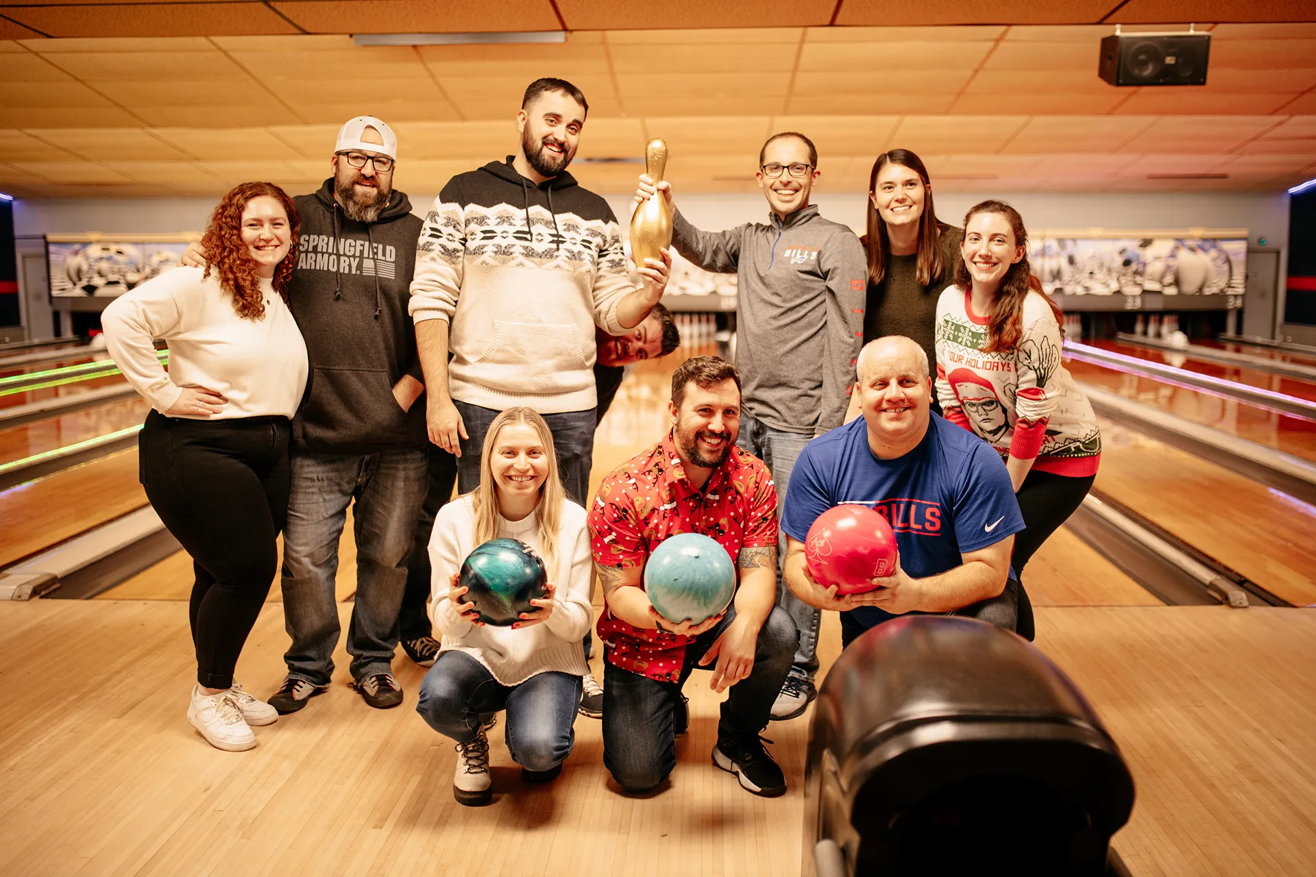 Group of nine smiling people posing in a bowling alley holding bowling balls and a golden pin.