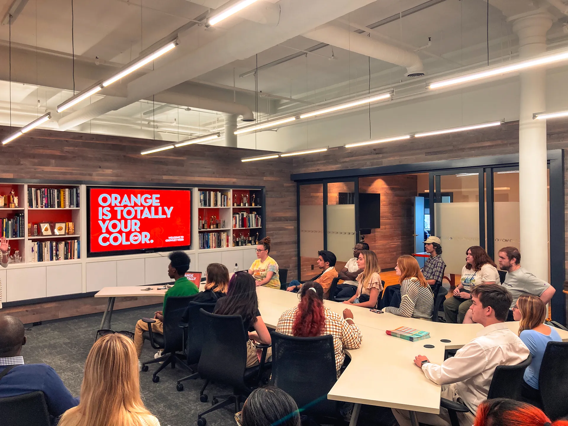 Group of diverse students seated around a conference table attentively watching a presentation screen that reads 'ORANGE IS TOTALLY YOUR COLOR.'