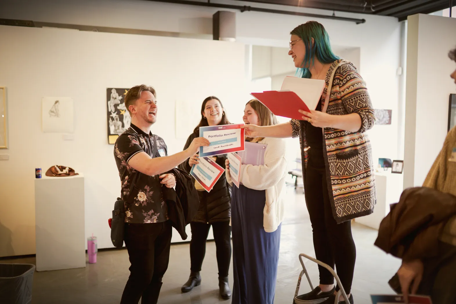 A group of young adults smiling and holding portfolio review certificates in a bright indoor space, one woman stands on a stool handing a certificate to a laughing man.